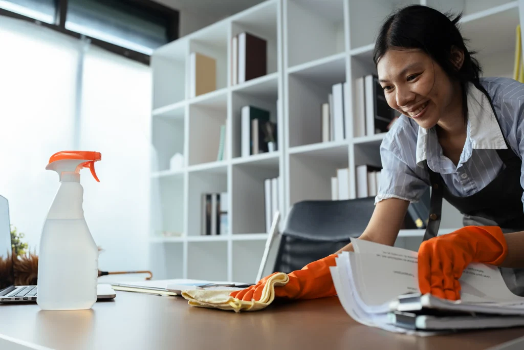 A person wearing orange cleaning gloves organizes documents on a desk, with a spray bottle and laptop nearby in a bright office setting.
